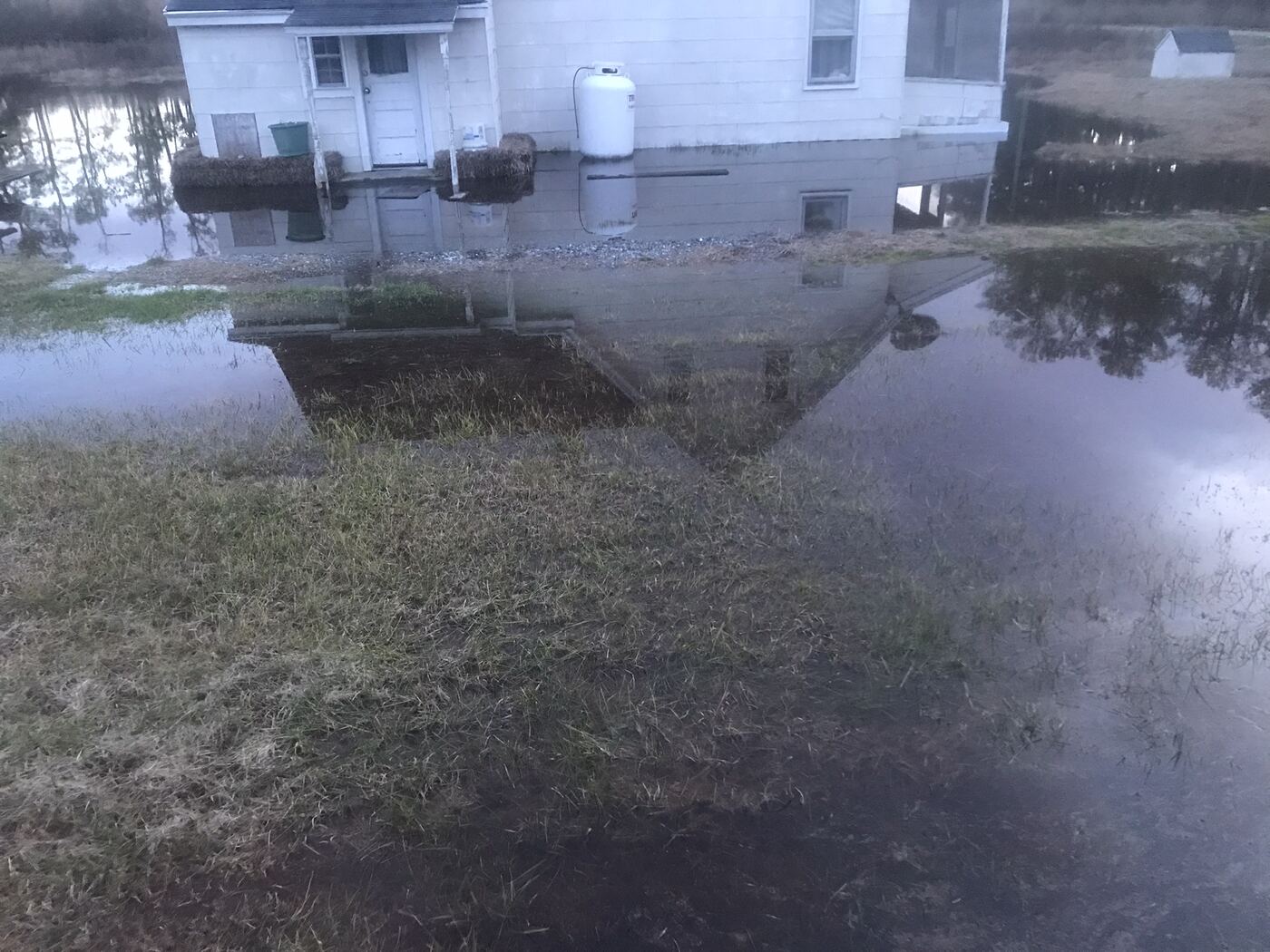Winter flood water spread across a yard with a white house and propane tank reflected in it